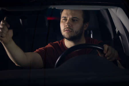 Handsome young millenial man driver sits at steering wheel of car at night adjusting up rearview...