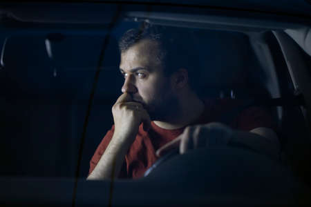 Handsome young man driver sits at steering wheel of car at night thoughtfully looking away....