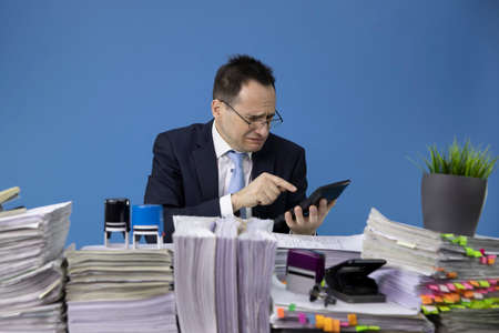worried weeping businessman considers losses on calculator sitting at table with piles of papers in...