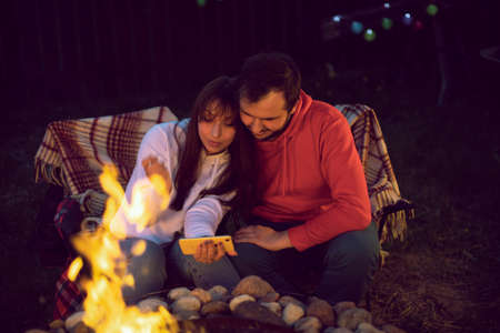 couple of lovers making selfies in glow of fire at romantic party by bonfire. Romantic camping couple sitting by bonfire in open air. Loving man and woman relaxing near campfire.の写真素材