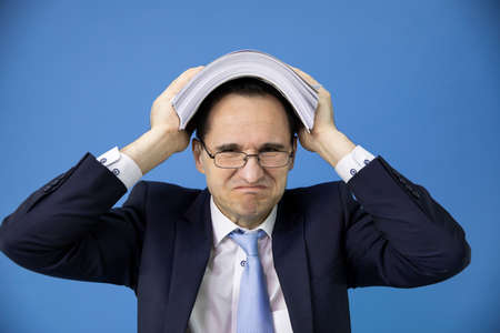 overworked overwhelmed office clerk covered his head with stack of documents. overhour job concept. Fatigue and overload concept. business stress and crisis concept. isolated on blueの写真素材