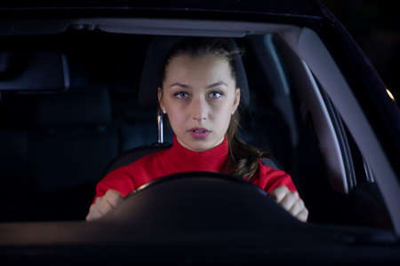Woman is driving car at night. Serious female driver sitting inside car looks confused forward to the road clings tight to wheel on night trip. Concept: nerves and transportation. Safe journeyの写真素材