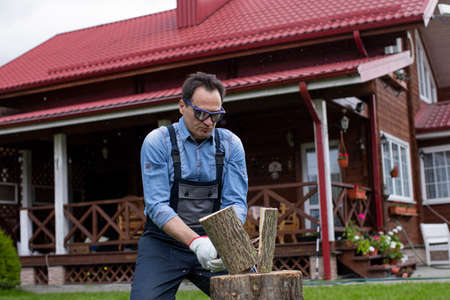 Strong man woodcutter in working uniform chopping wood with sharp ax on wooden hemp, sawdust fly to...