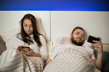 Couple in bed. Brunette girl and bearded guy looking at their smartphones. concept of loneliness in...
