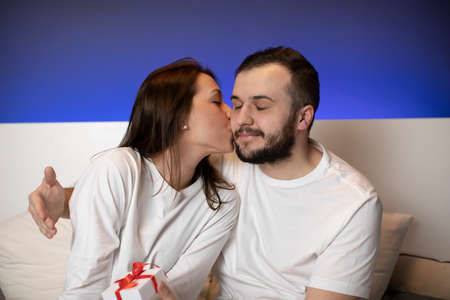 couple of lovers wearing white pajamas in bed. Beautiful Brunette girl kissing her happy bearded...