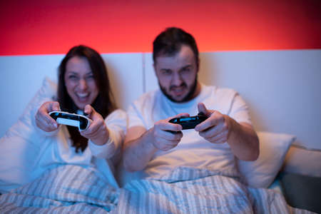 couple of gamers in love passionately playing video games in bed with joysticks. Selective Focus on game console remote controls. computer addiction concept. Common hobbies, shared valuesの写真素材