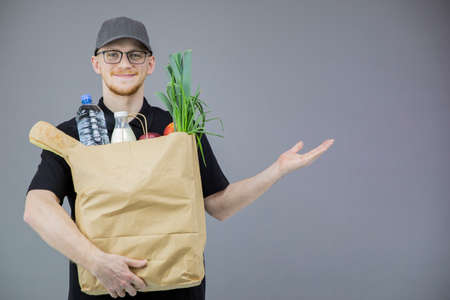 young Handsome Caucasian delivery man in black uniform handling paper bag of food smiling to camera pointing hand at your offer isolated on gray. express grocery delivery service concept copy spaceの写真素材