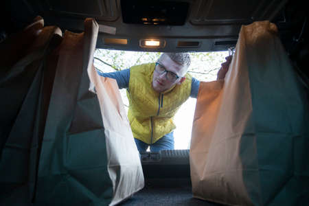 Handsome Caucasian guy in glasses delivery man putting Shopping paper bags into car trunk. courier...