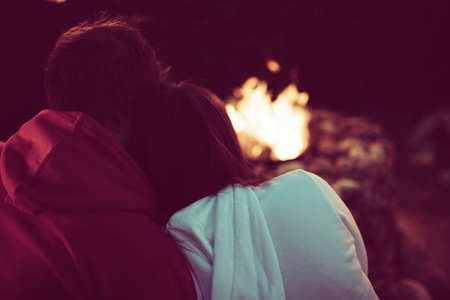 Portrait of a loving married couple sitting by the fire in nature. A man hugs a woman, view from the back. Rest in the country with a camper. Family holidays away from city noise. Toned sepiaの写真素材