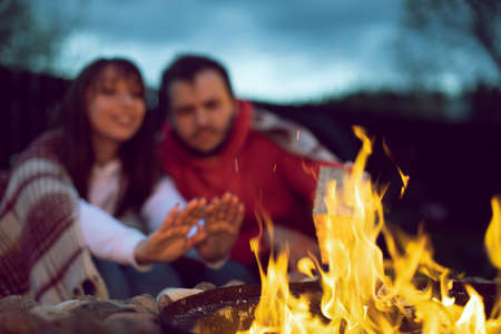 Portrait of happy lovely couple sitting by fire. Woman warms her hands by the fire. Relaxing near the fire in the nature concept. Family holidays away from city noiseの写真素材