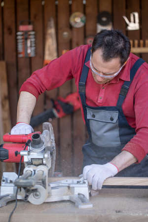 A Caucasian man in protective overalls and glasses cuts a wooden oak plank with a circular saw....