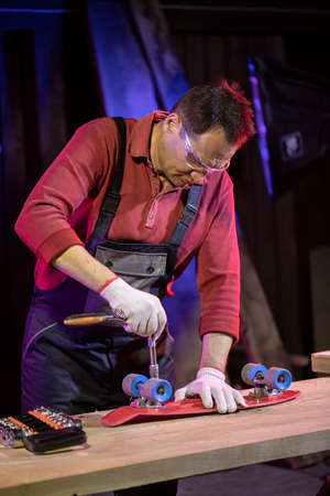Mature middle-aged man in jumpsuit fixing red plastic kids skateboard in home workshop with socket...