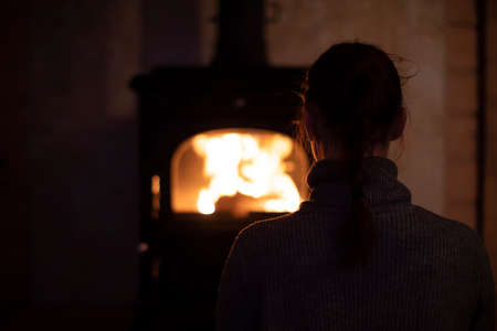 A young introvert girl sits at home by the fireplace in the evening and watches the fire. Reading books in the evenings at home. Cozy home interior of a wooden house. Relax after a hard day.の写真素材