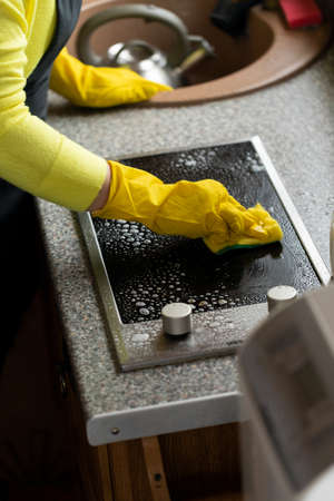 Close up person in yellow rubber gloves cleaning house wipes kitchen worktop, washes induction stove with sponge. Housework cleaning service, household chores, quick easy kitchen cleaning productsの写真素材