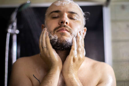 Close up view of handsome man with closed eyes washing beard in bathroom, taking shower. Lifestyle, beauty, personal care, relaxing time, young guy enjoying showering, Body, skin, hair hygieneの写真素材