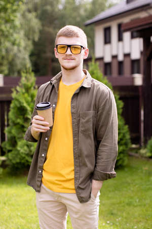 Portrait of young hipster guy businessman freelancer standing outdoors, resting in park with cup of...