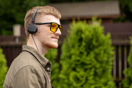 Pensive young handsome man in headphones standing outdoors, listening music educational podcast...