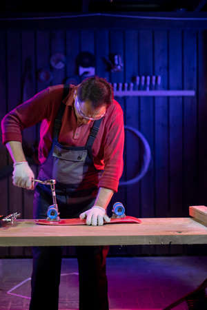 Mature middle-aged man in jumpsuit fixing red plastic kids skateboard in carpentry with socket...