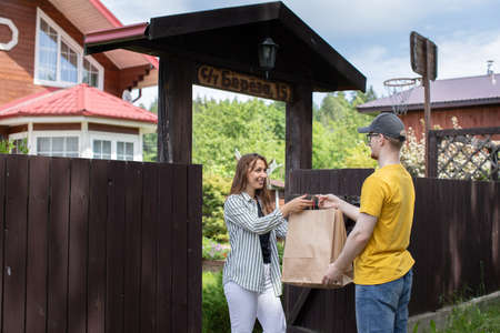 Beautiful woman customer receiving order from courier, Delivery man in yellow t-shirt handing paper bags to recipient in front of country house, delivery service concept, e-commerce, online shoppingの写真素材