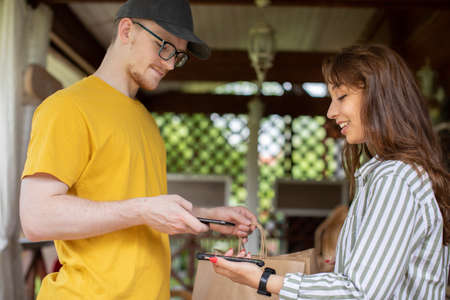 Smiling young delivery man holding paper bags while beautiful young woman makes cashless payment for...