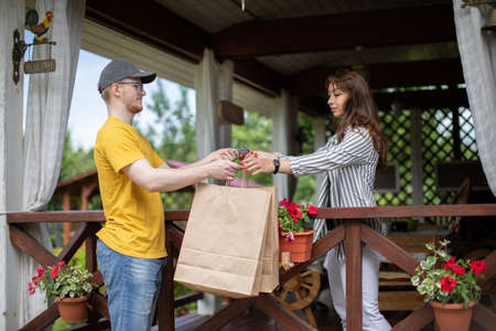 Delivery man handing paper bags to female recipient, beautiful young woman customer receiving order from courier at home, pays for order in cash, express delivery service, online shopping conceptの写真素材