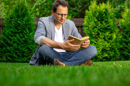 Adult handsome man sitting on grass in park with paper book resting relaxing, male businessman freelancer reading outdoors. Summer, education, study, training courses and programs concept. Copy spaceの写真素材