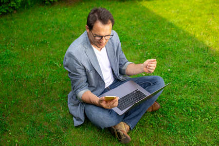 Middle aged businessman works with laptop and mobile phone outdoors, sits on grass in park, happy...