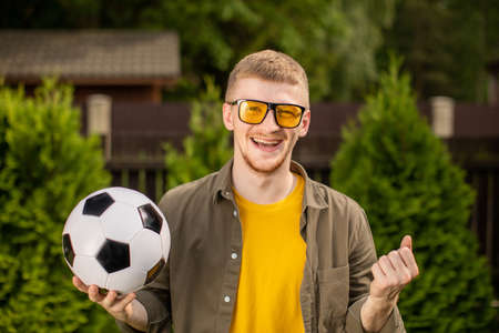 Excited hipster guy holding soccer ball smiling and screaming, Happy male football fan support sport team, celebrating victory. People emotions, sports, entertainment concept, successful bet on sportsの写真素材