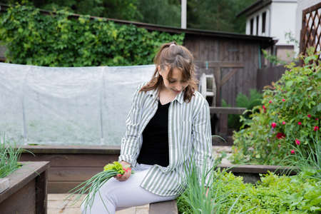 Young beautiful woman gardener sitting on raised garden bed harvests fresh herbs green leek and...