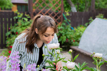 Attractive young woman gardener smelling fragrant white flowers of blooming peony in outdoor flower bed with eyes closed and smile, home garden, summer spring time, beauty, aroma, happy life conceptの写真素材