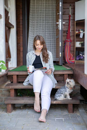 Young beautiful woman resting outdoors with cup of coffee, reading morning press on porch of country house, housewife chilling outside with dog. Summer lifestyle, leisure free time concept. Copy spaceの写真素材