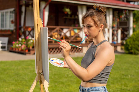 Young beautiful woman female artist painting on easel outside on loan near country house, holding paint brush in mouth, outdoors relaxing activity, creative hobby, creation, inspiration conceptの写真素材