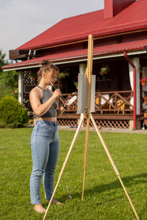 attractive girl paints easel painting at her country house outdoor. Pretty woman is painting. Open air session. Outdoor time for hobbiesの写真素材