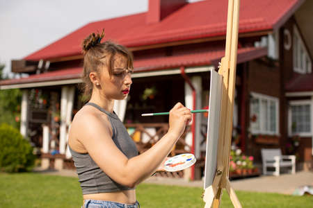 Young beautiful woman female artist painting picture on easel outside on loan near country house,...