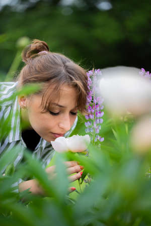 Attractive sexy young brunette woman sniffing white pink peony amidst flowers of lupine on green...