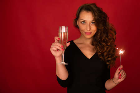 Young successful woman with champagne and sparkler looks at camera, red background, copy space. New Year Christmas holiday congratulation, copy space. Achievement, business awards, reward conceptの写真素材