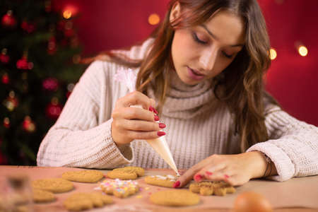 Christmas bakery. cute brunette girl icing Christmas gingerbread with icing-sugar with Christmas tree and red Xmas decoration on blurred background.の写真素材