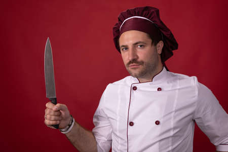 Unshaven handsome male chef cook holding chefs knife looking at camera isolated on red background. professional fatigue, rest time, Red danger level. Chief knife - common murder weapon, stabbingの写真素材