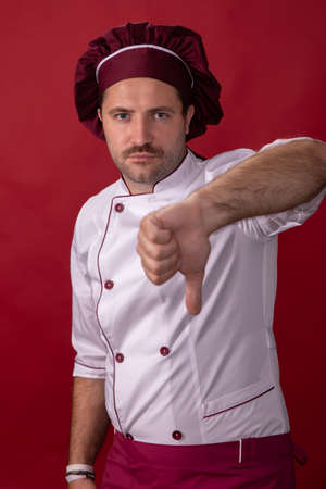 Unshaven handsome male chef cook shows thumb down looking at camera isolated on red backdrop. Chef dislike gesture. Expired products, low level, no culinary ability, bad food, disgusting taste conceptの写真素材