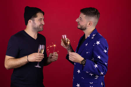 Two attractive stylish gay men celebrating New Year party, homosexual couple holding sparklers and glasses with champagne congratulating each other on red background. Romantic Christmas conceptの写真素材