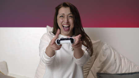 Joyful young attractive woman in white pajamas playing video games with joystick in hands. Sexy...