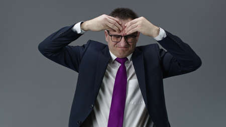 Tired and puzzled handsome middle aged businessman in formal clothes, tie and glasses suffering from headache rubbing forehead with hands, isolated gray background, deadline, stress condition conceptの写真素材