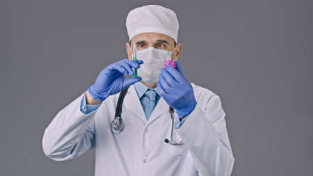Male middle aged doctor in medical uniform and mask holds syringe, injects test vaccine in covid-19...
