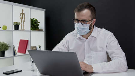 Attractive young businessman in white shirt, glasses and protective mask working away from office (telecommuting) at laptop during quarantine due to coronavirus global pandemic, self isolation at homeの写真素材