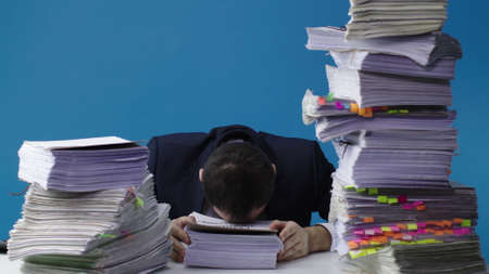 Stressed businessman accountant manager takes stack of unfinished documents from large pile of office papers, in despair lays head on table. Stress at work, overworked, bureaucracy, paperwork conceptの写真素材