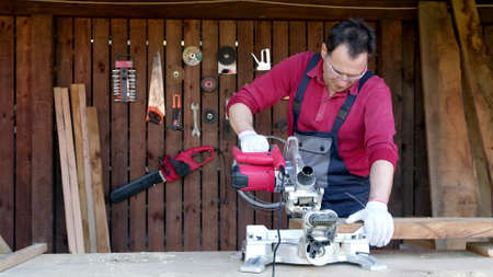 Male joiner processes wood bar using a miter saw in a workshop. Work with miter saw for cutting boards and bars. Home renovation with professional construction and repair tool.の写真素材
