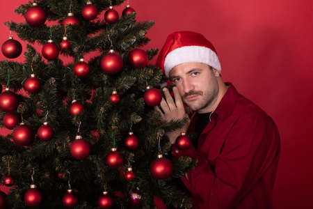 Young man with mustache in Santa hat gently hugs nestles against Christmas tree on red background, copy text space. Choosing best xmas spruce fir, waiting for New Year holiday offers discounts conceptの写真素材