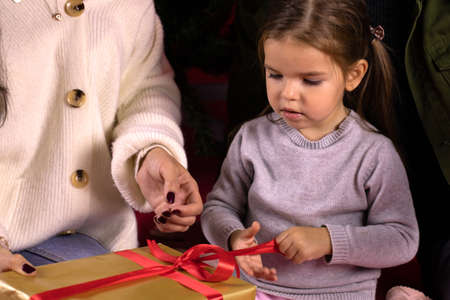 Toddler girl with her parents opens Christmas gift sitting under Christmas tree close up portrait. Mother helping his daughter to unleash red bow on gift box. Family traditions conceptの写真素材