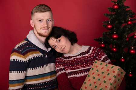 Lovely cute couple posing at home in front of xmas tree, celebrating New Year, calm young man and beautiful woman with Christmas gift box on red background. Winter family holidays, conceptの写真素材