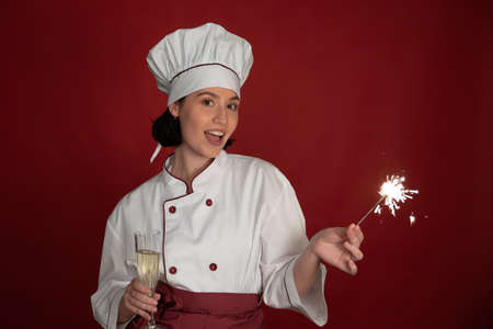 Young attractive sexy female chef looking at camera with glass of champagne and Burning sparklers in her hands on red. Young cheerful girl Cook celebrating Christmas and New Year Eve in festive moodの写真素材